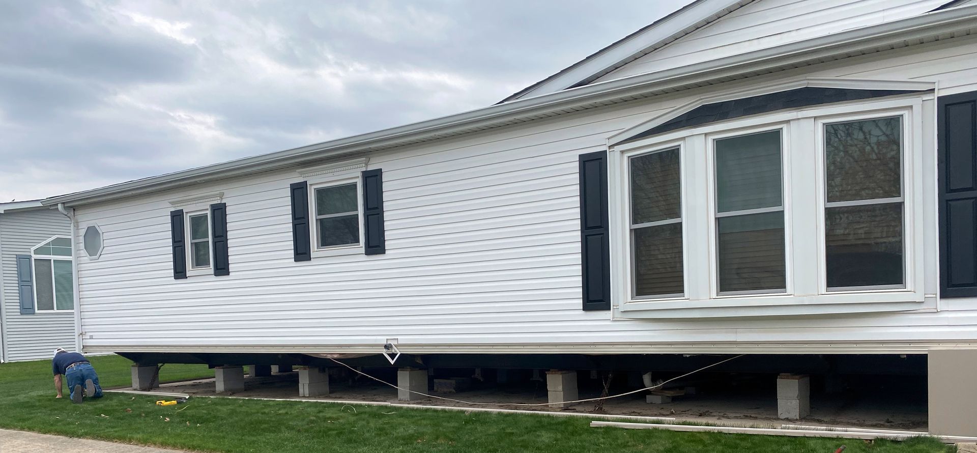 White mobile home on blocks with black-trimmed bay window, seen from the side on a grassy lot