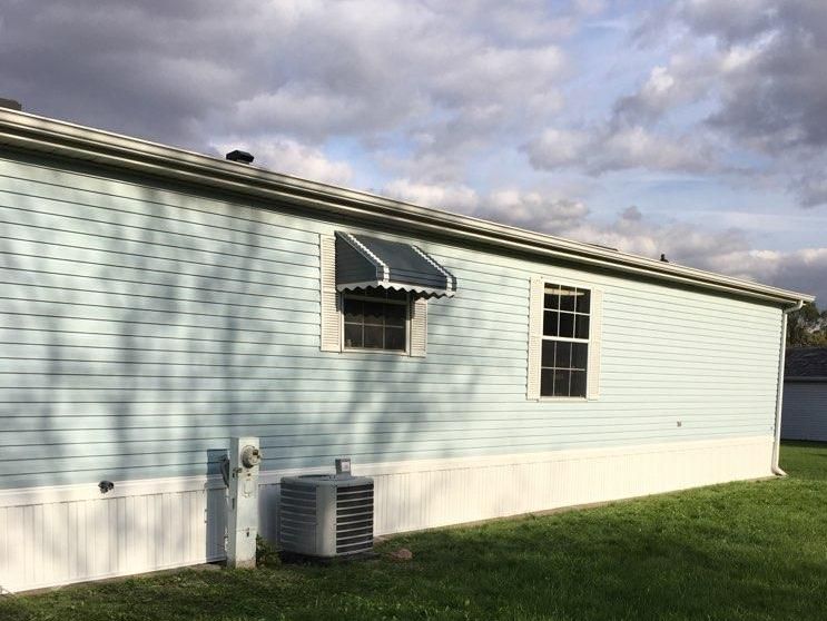 Exterior of a light blue mobile home with two windows, an awning, and an air conditioner unit on the side
