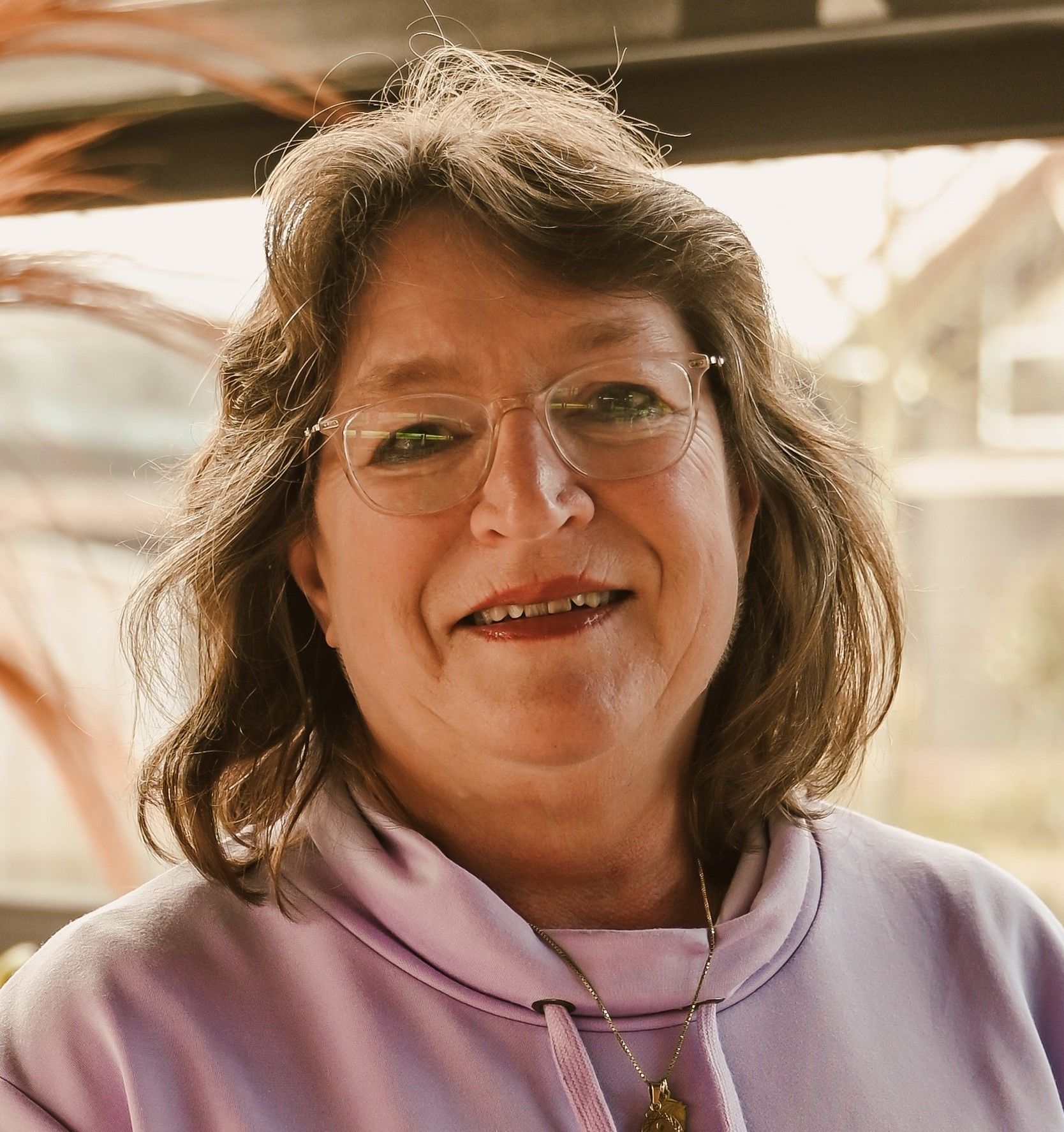 Vrouw met bril lacht; lichtpaarse hoodie, binnenomgeving.