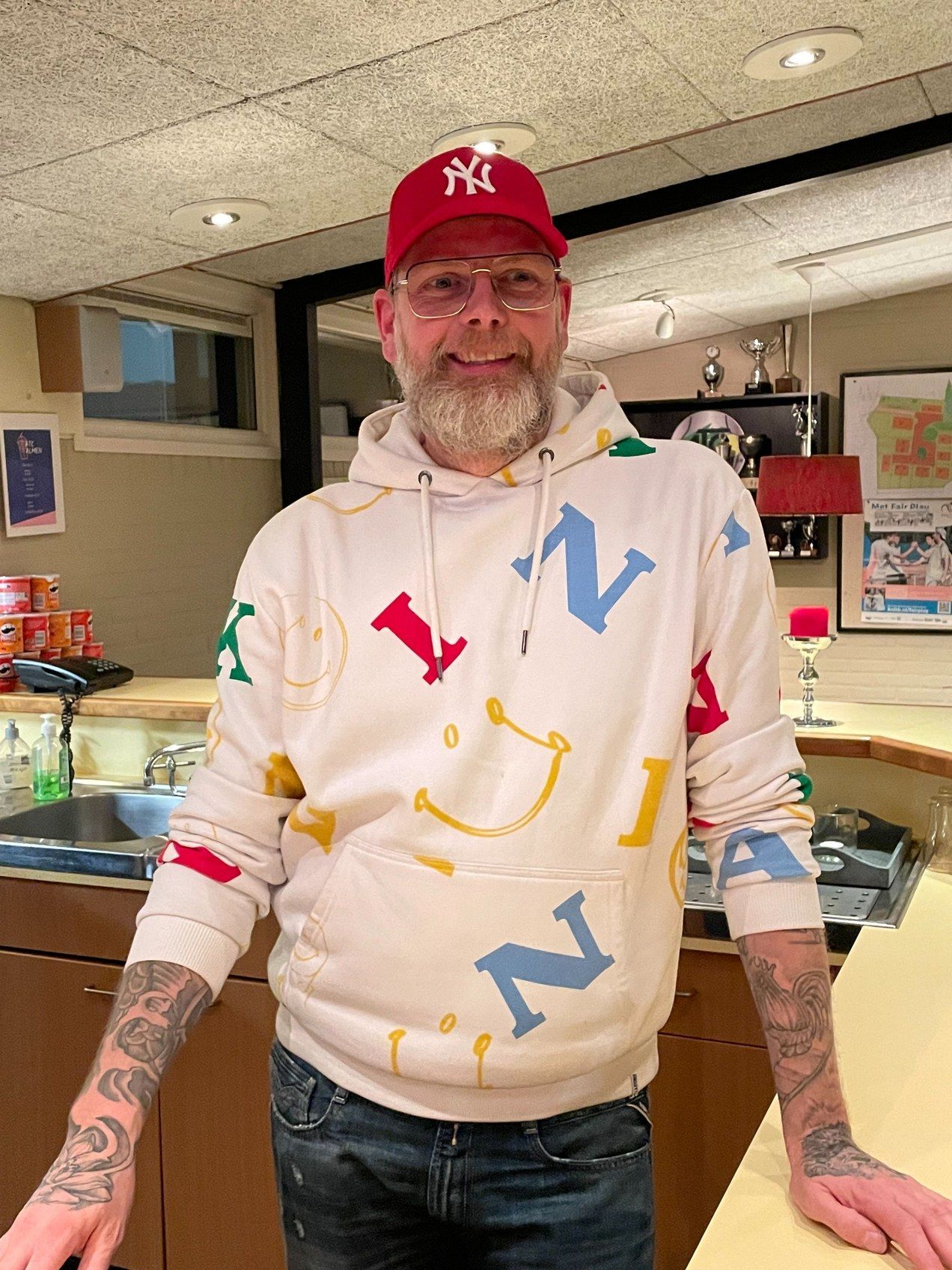 Een man met een rode Yankees-pet en een witte hoodie met kleurrijke letters staat binnen bij een toonbank.