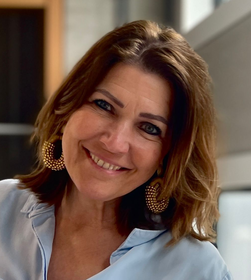 Vrouw met schouderlang bruin haar lacht, draagt ​​gouden oorringen en een lichtblauwe blouse.