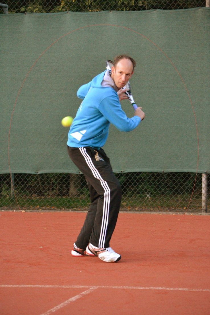 Een man in een blauwe hoodie zwaait met een tennisracket en de bal in de lucht op een rode gravelbaan.