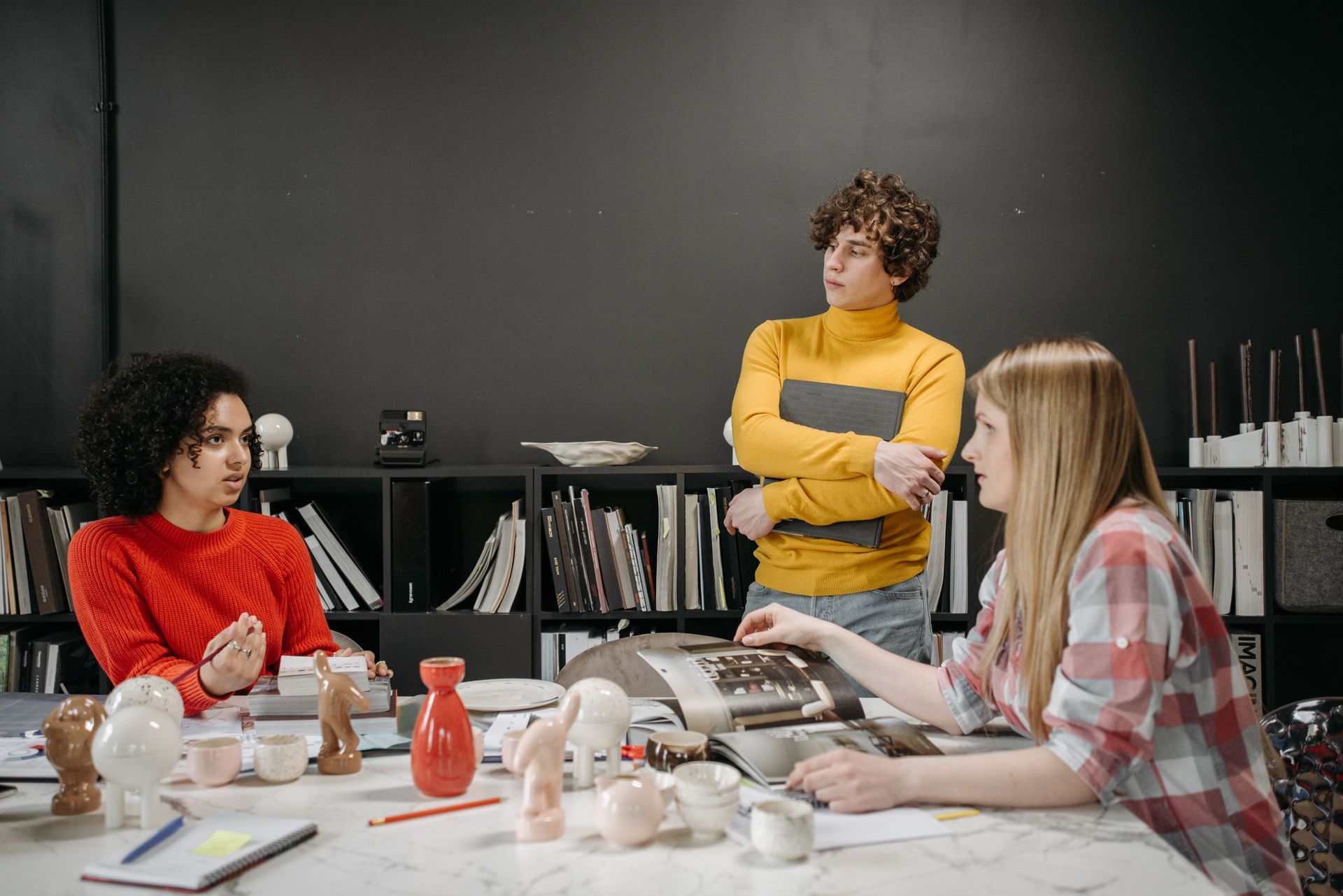 Three people collaborating at a table in a classroom or studio, with art supplies and shelves in the background.