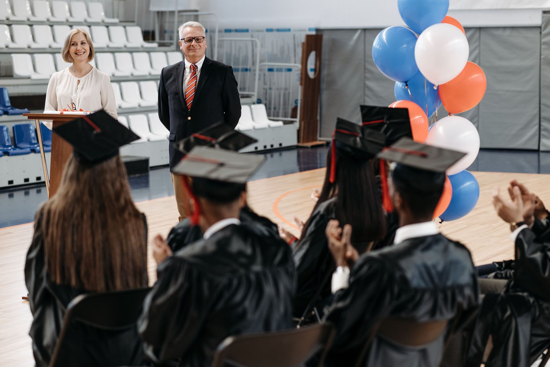 Graduates in caps and gowns applaud two speakers on a gymnasium stage with red, white, and blue balloons.