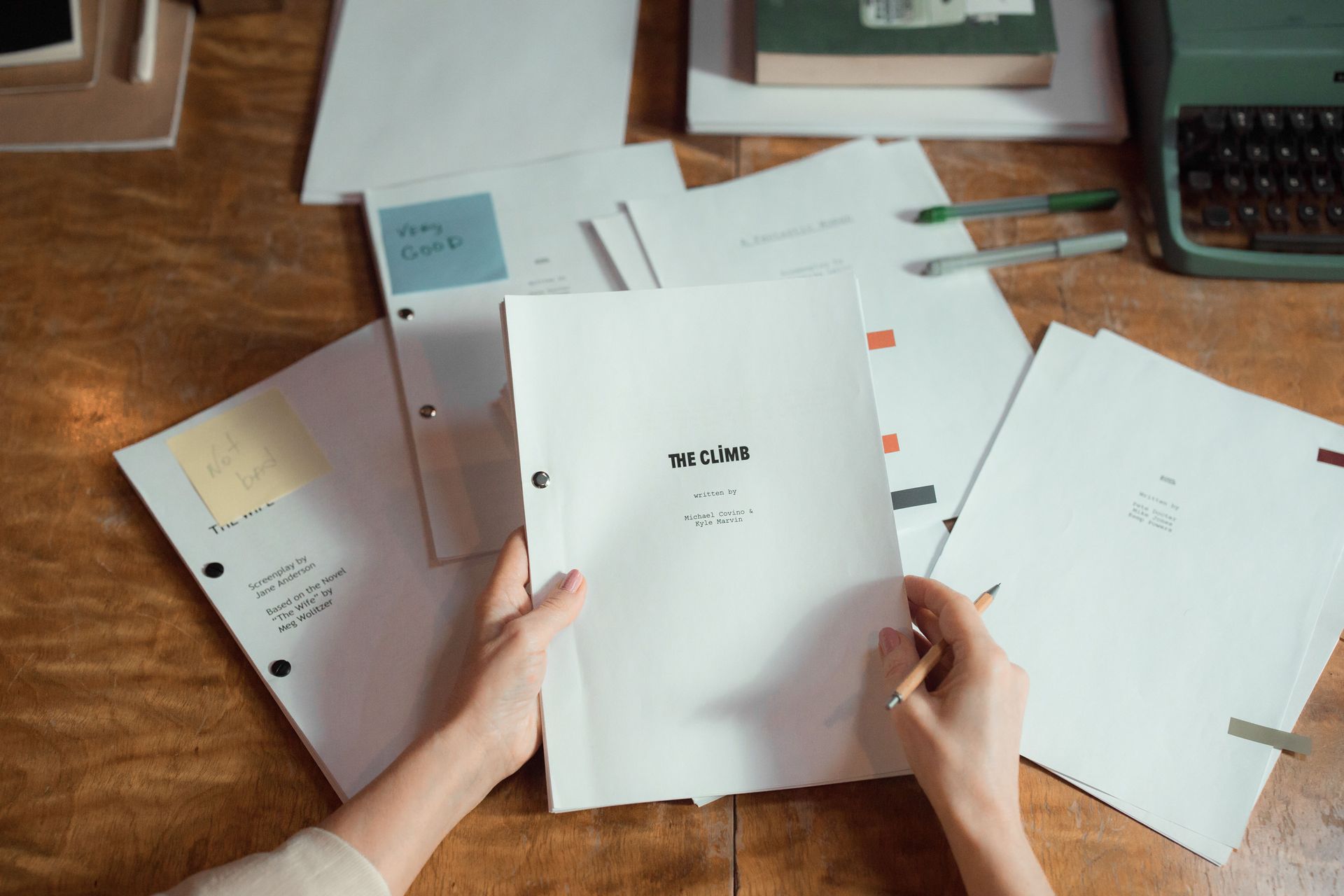 Hands holding a white booklet over scattered planner pages on a wooden desk with a laptop and pencil case nearby