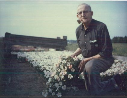 A man is kneeling down in a field of flowers with a pipe in his mouth