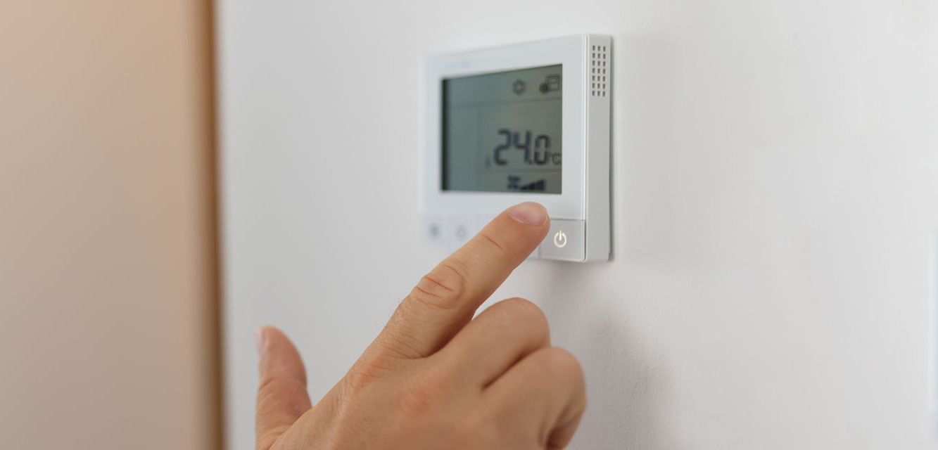 Le doigt d'une personne appuie sur un bouton d'un thermostat blanc ; l'écran affiche 24,0.