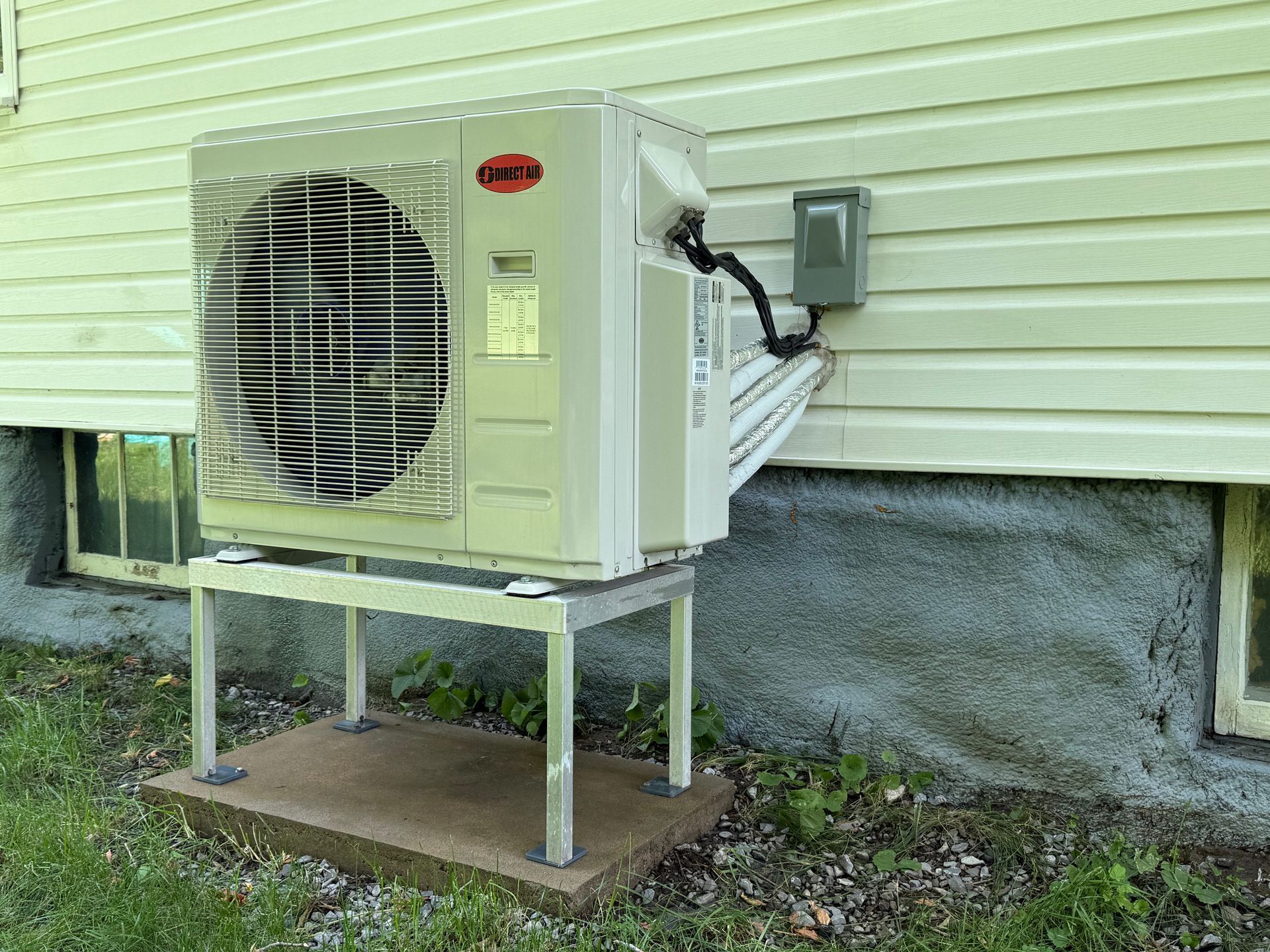 Unité de climatisation sur support, montée sur un mur extérieur de maison à côté d'une fenêtre.