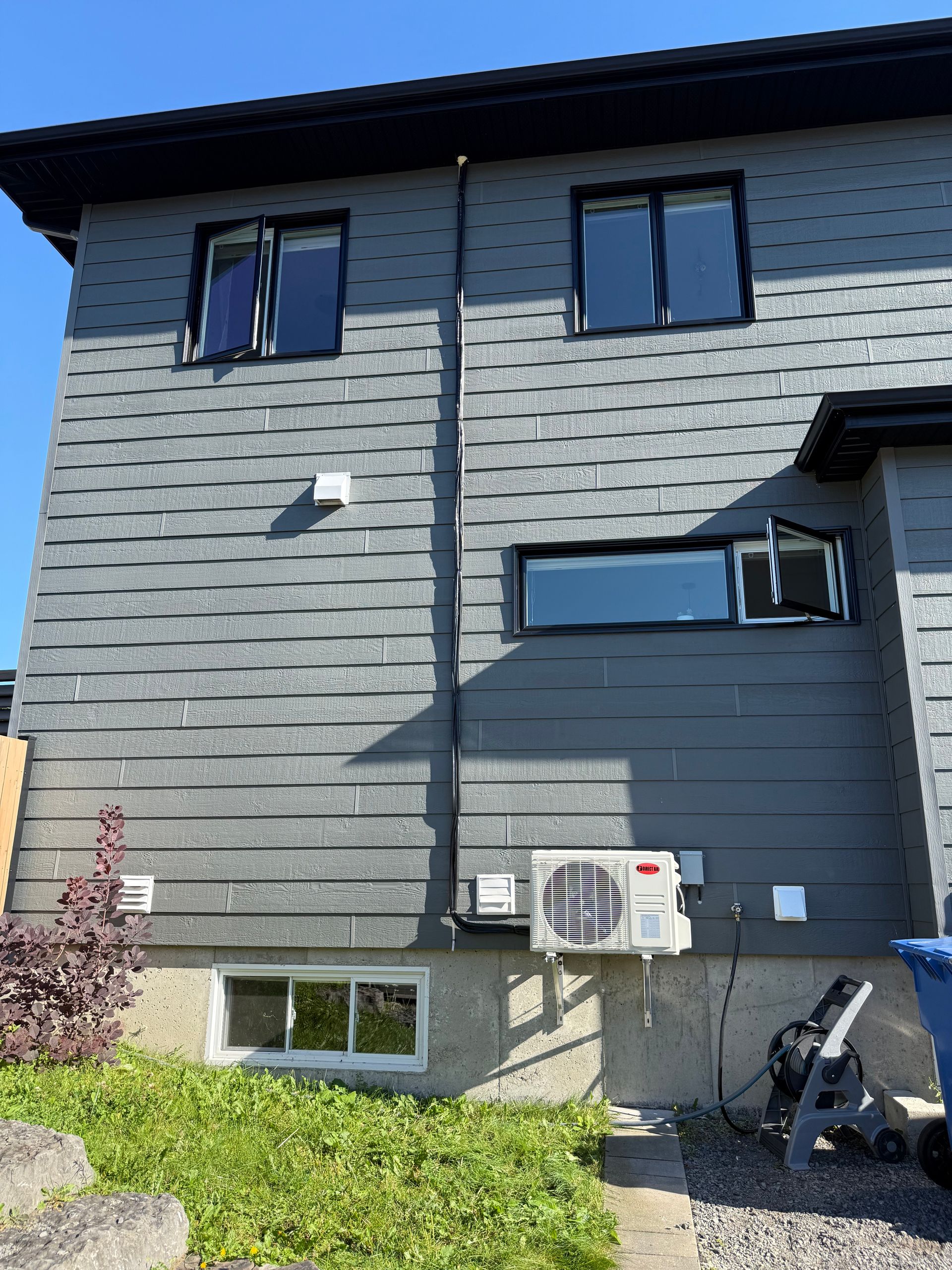 Extérieur d'une maison grise avec fenêtres, climatiseur et conduits électriques. Feuillage vert au premier plan.