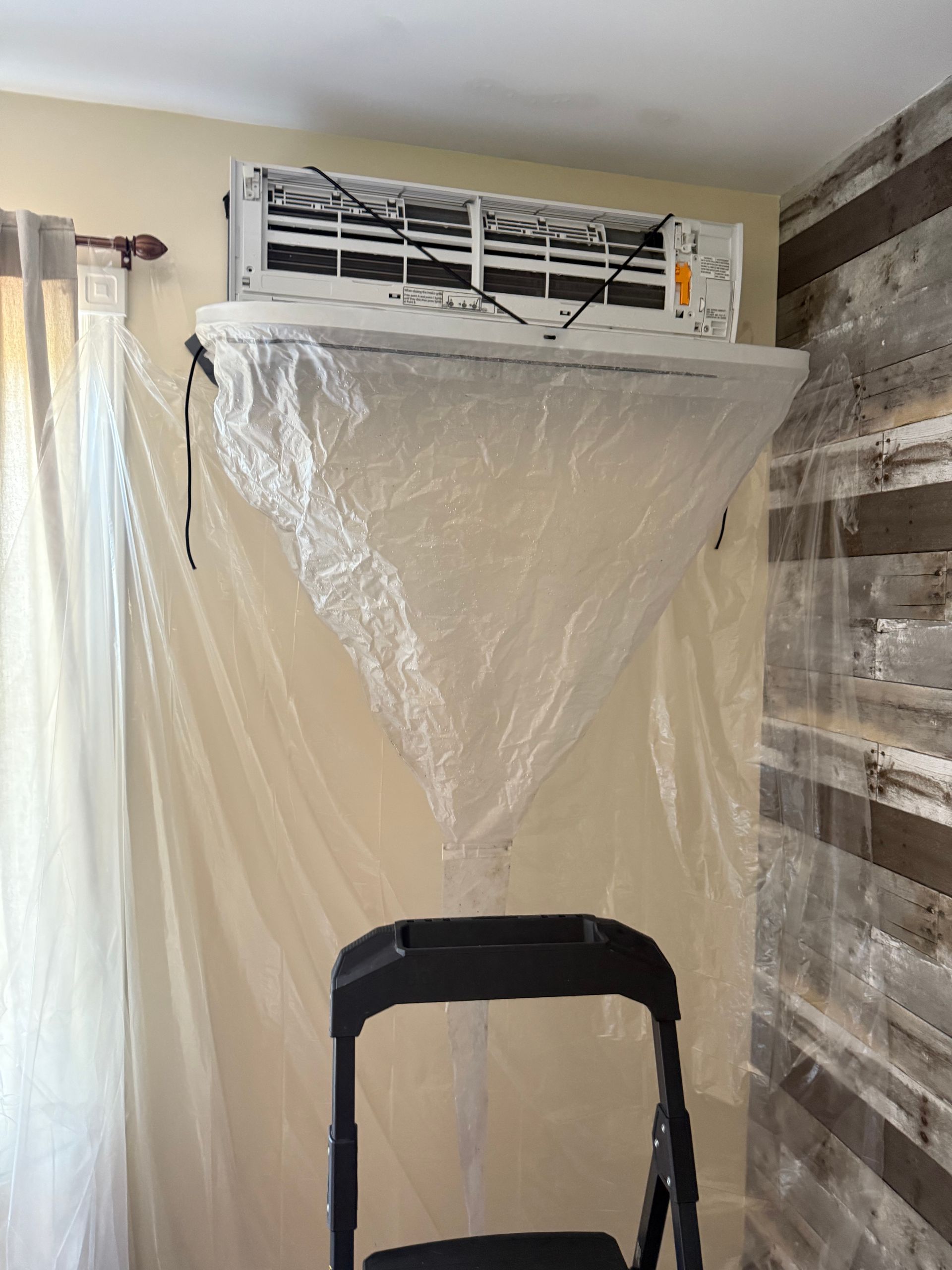 Climatiseur recouvert de plastique, au-dessus d'un marchepied noir, contre un mur lambrissé.