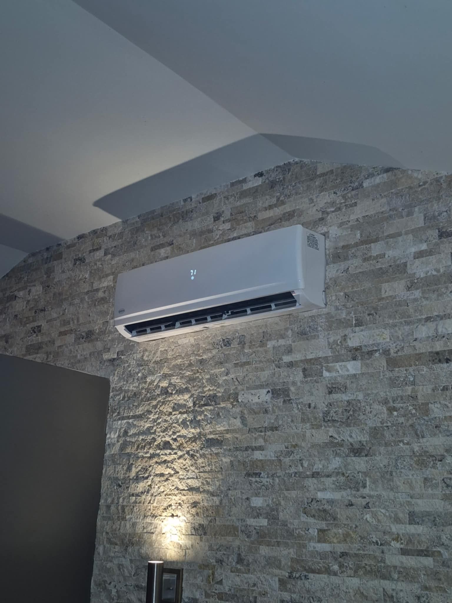 Climatiseur monté sur un mur en pierre texturée. Un spot en dessous éclaire le mur.