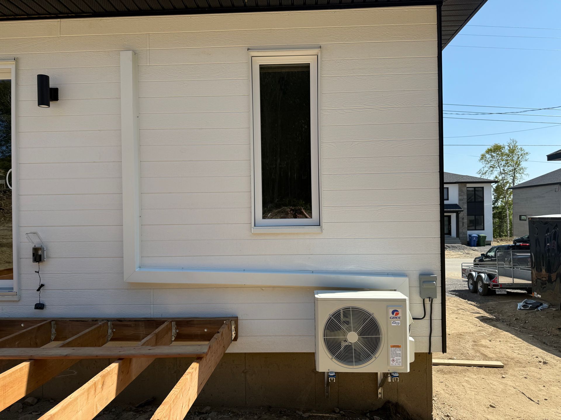Extérieur d'une maison blanche avec une petite unité de climatisation installée sous une fenêtre étroite, avec une terrasse partiellement construite.