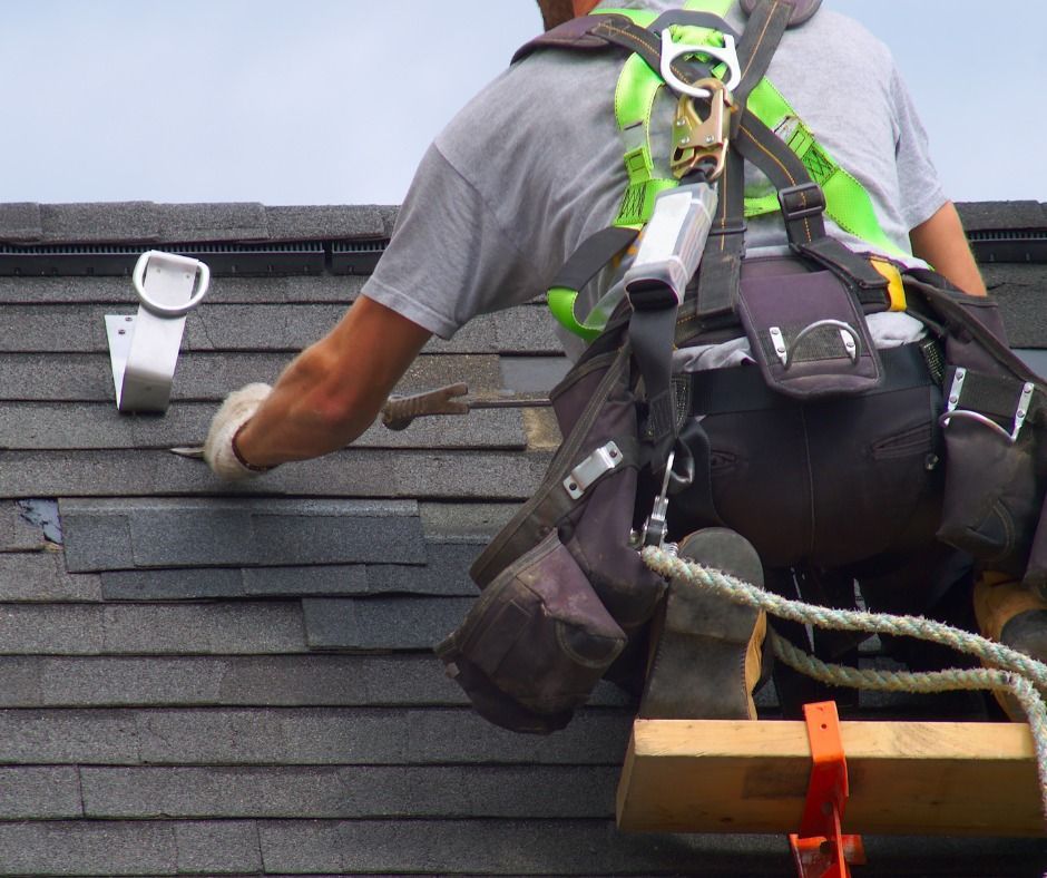 Worker in safety harness repairing a shingled roof with a tool bag and ladder.