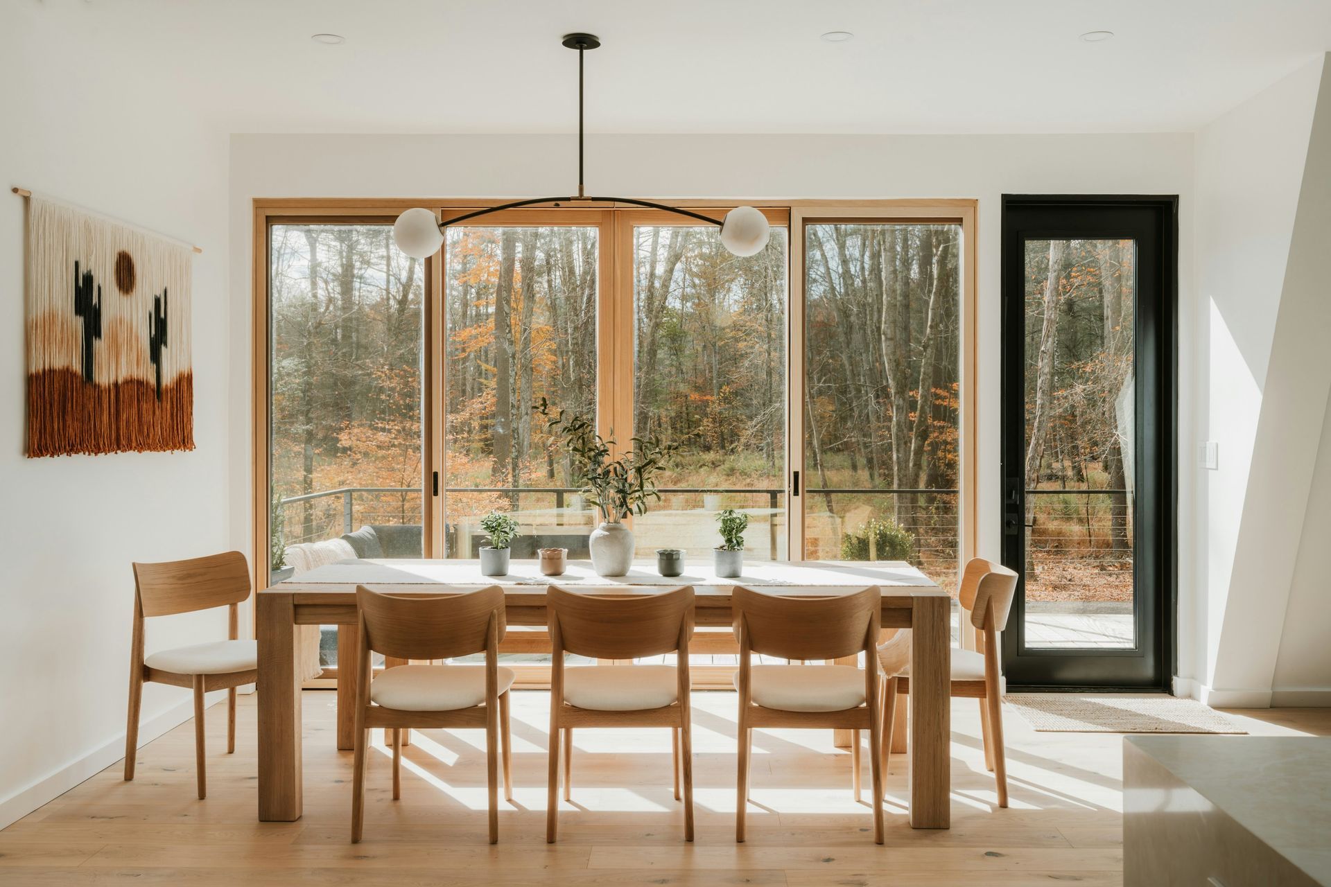 Bright dining room with wooden table and chairs beside large windows and a glass door overlooking trees