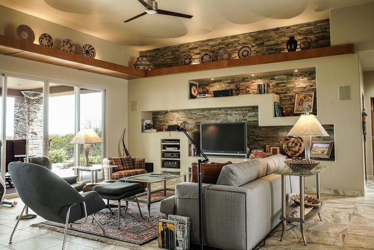Bright living room with a gray sofa, swivel chair, fireplace, and large TV beneath a wooden mantel.