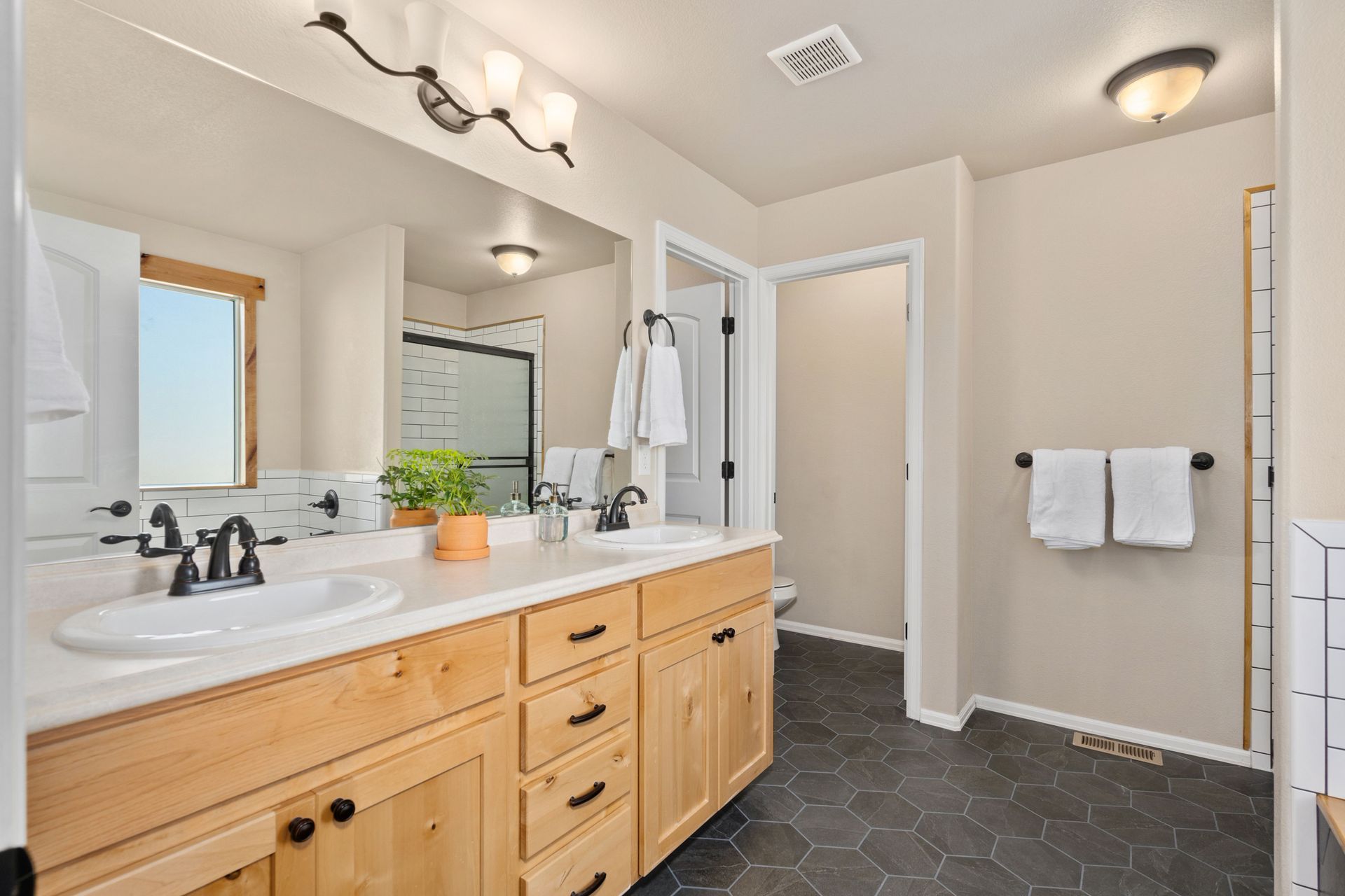 Bright bathroom with double vanity, large mirror, white walls, and dark hex tile floor.