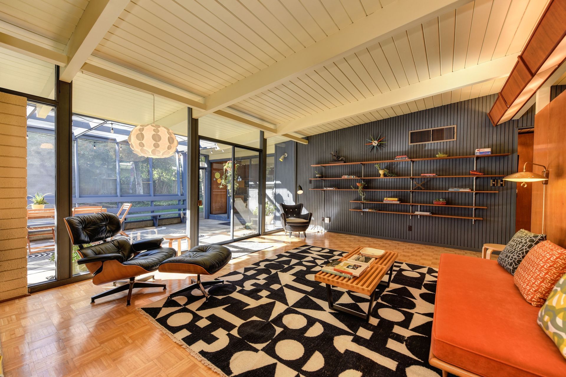 Modern living room with orange sofa, patterned rug, shelving wall, and large windows overlooking outdoors