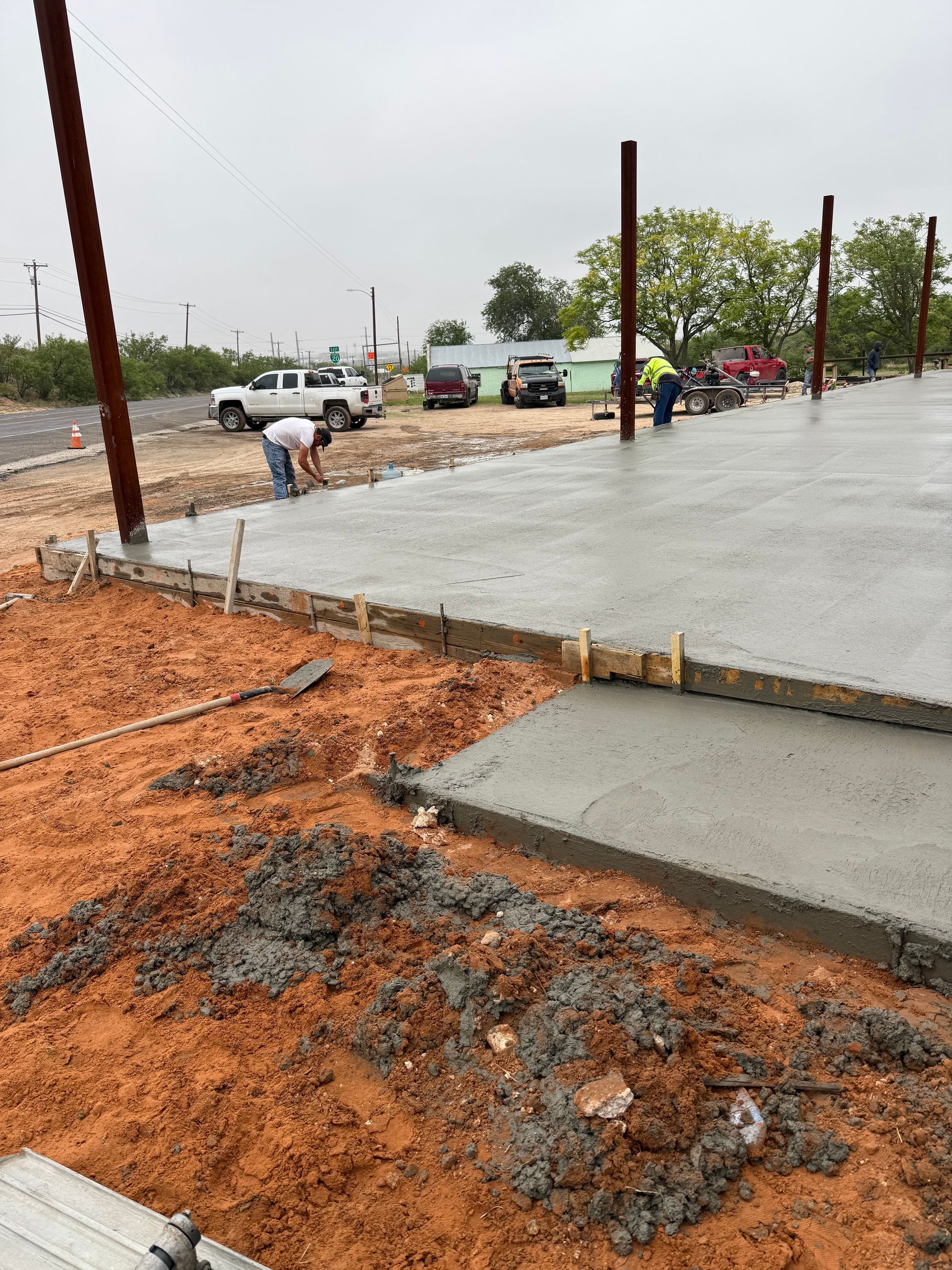 Construction workers pouring a concrete slab beside orange dirt and rebar on a rainy roadside