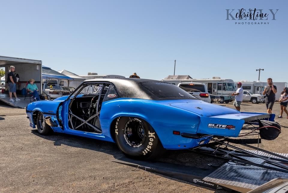 Blue drag racing car on a trailer, prepared for competition. Sunny outdoor setting.