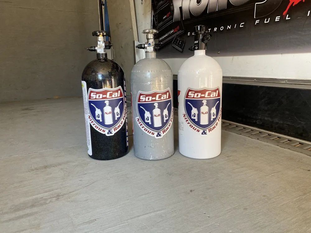 Three nitrous oxide tanks with So-Cal Nitrous & Fuel stickers: black, gray, white, on a concrete surface.