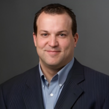 Headshot of a smiling person wearing a blue collared shirt and navy pinstripe blazer against a solid gray background.
