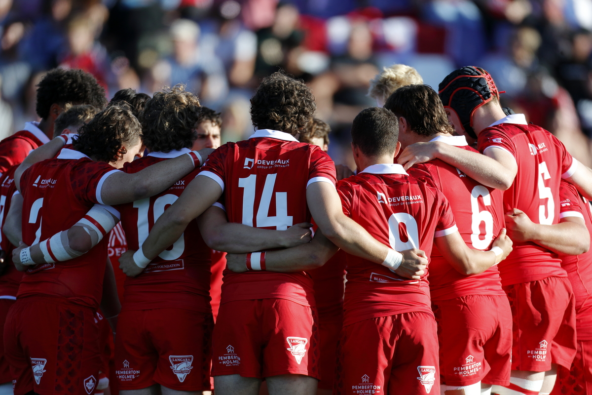 A rugby team in red uniforms stands in a huddle on a field, arms linked and facing inward, with spectators in the background.