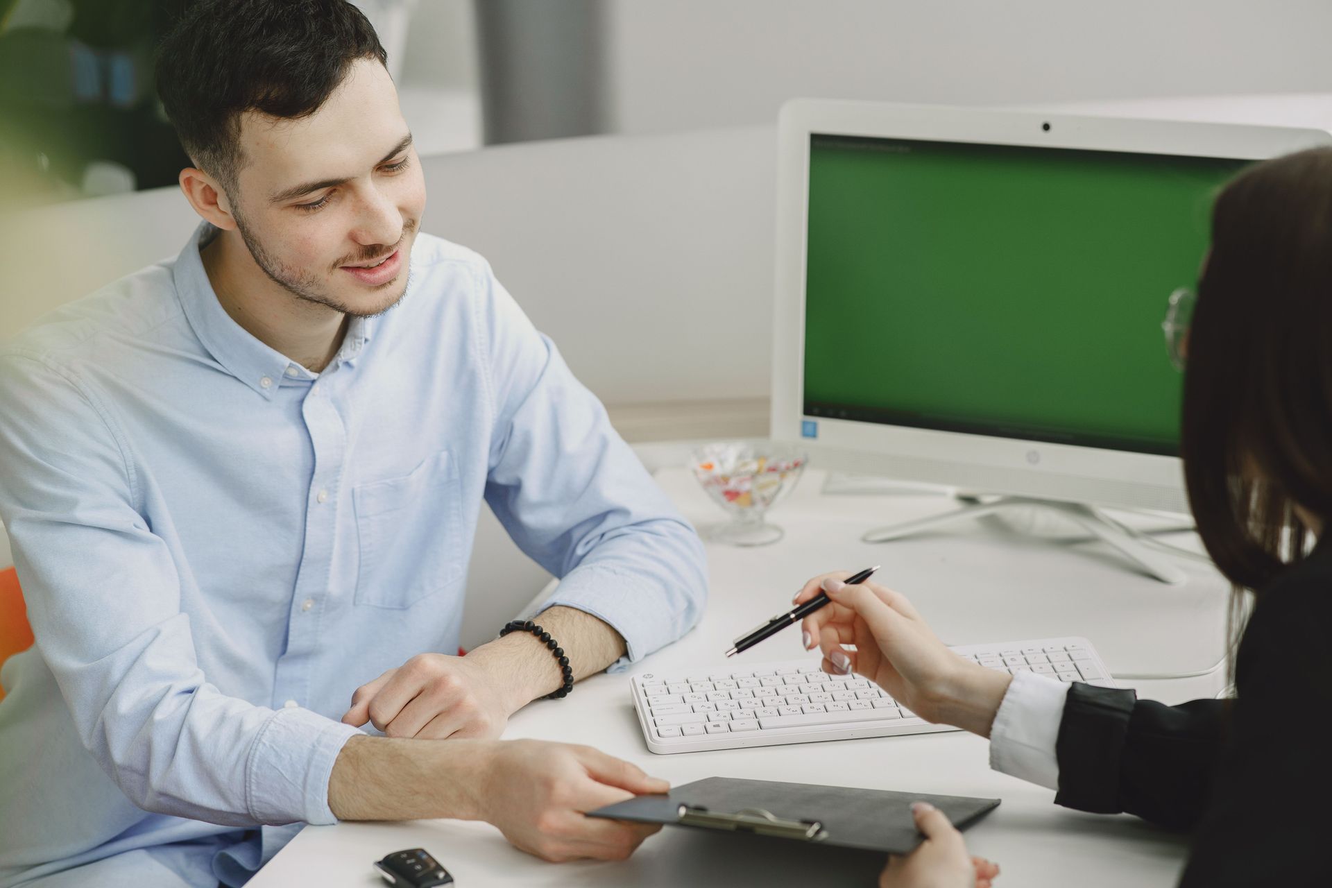 Two people discuss a document at a desk, with a laptop and green monitor in the background.