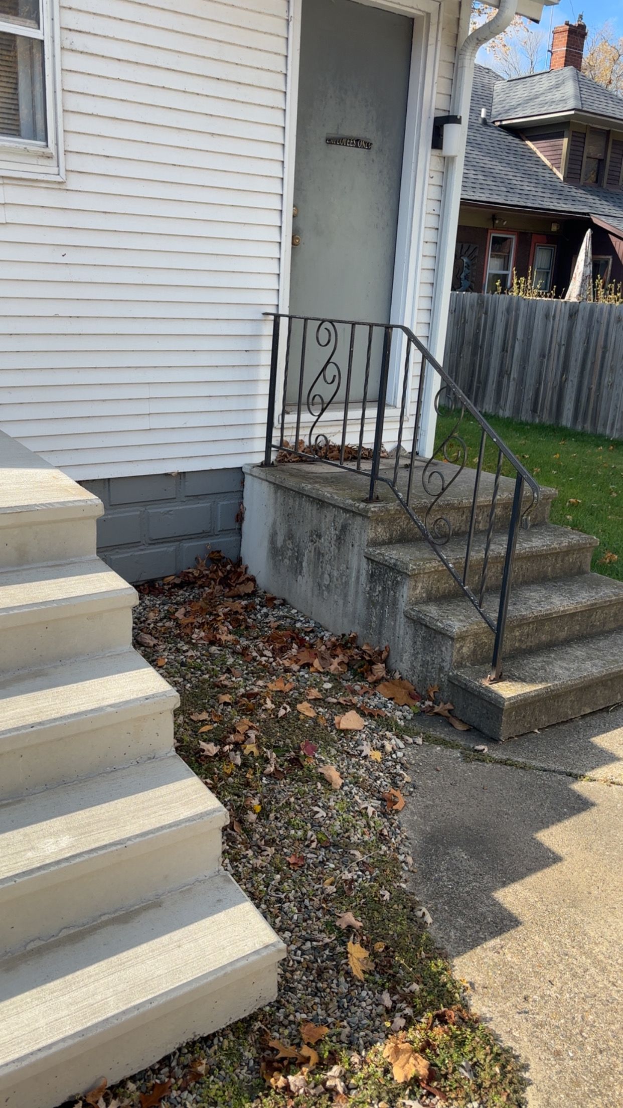 Concrete steps leading up to a door with a black iron handrail. Dry leaves on the ground.