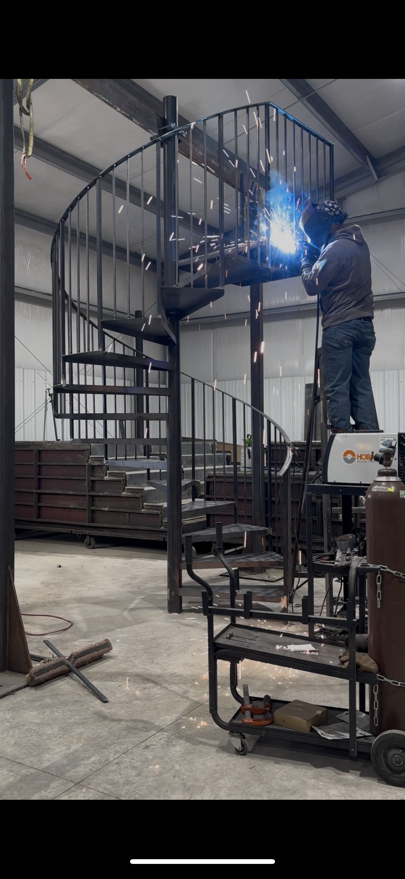 A worker welding a metal spiral staircase in a workshop. Sparks fly from the welding torch.