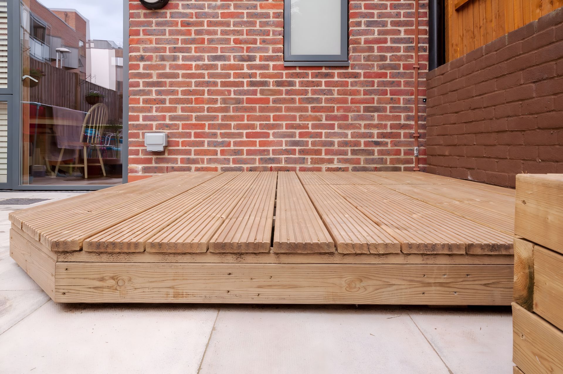 Wooden deck on a patio, with brick wall and glass door in background.