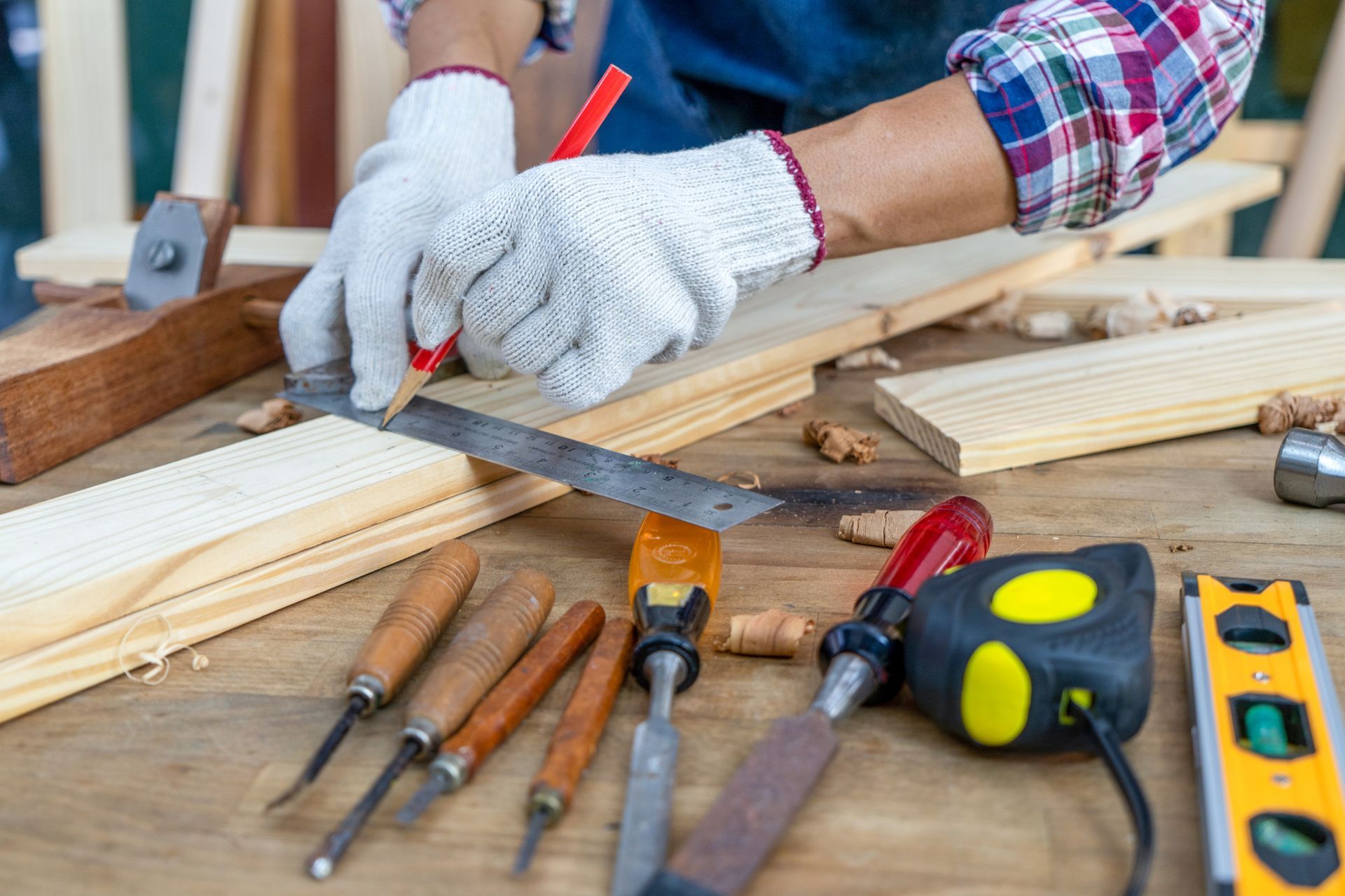 Carpenter wearing gloves marking wood with a pencil and ruler; tools on a wooden workbench.