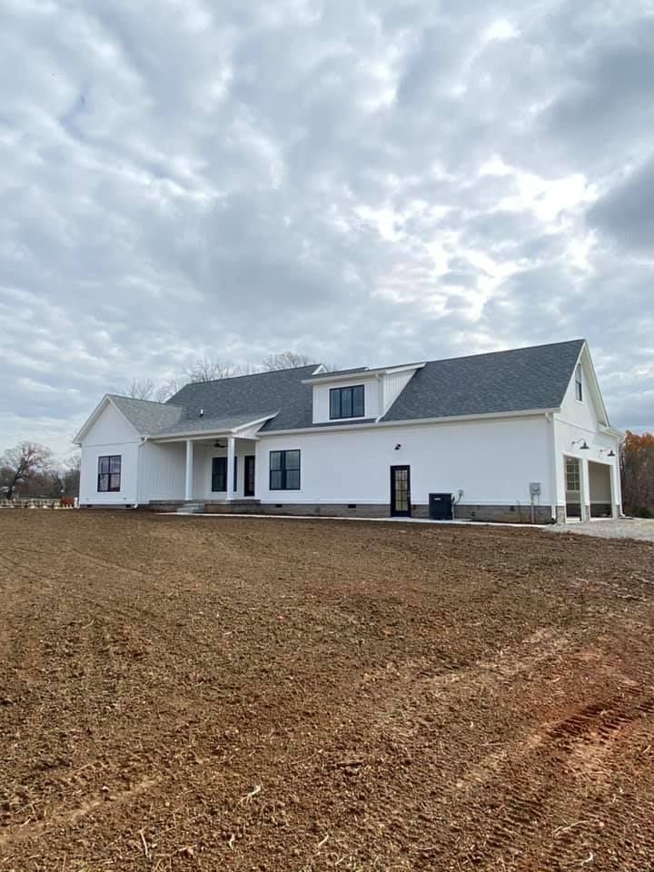 A newly built white house with a gray roof and a front-facing garage, situated on a large, cleared dirt lot under clouds.