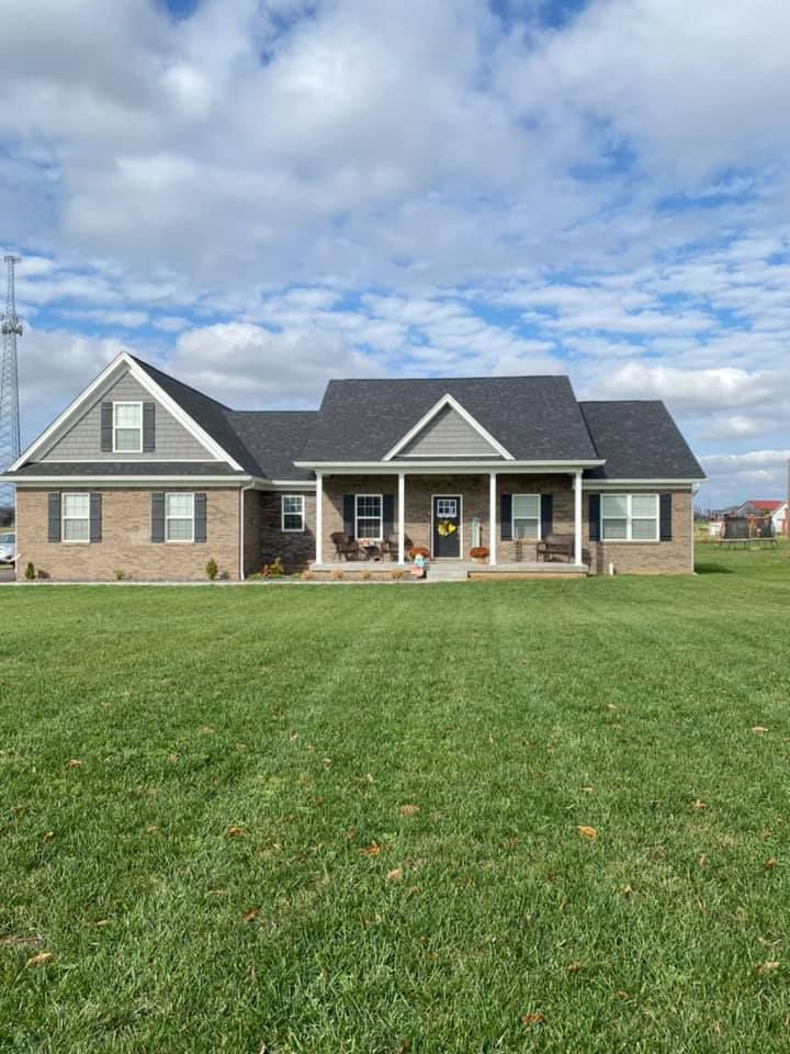 A single-story, tan brick suburban house with a dark grey roof and a front porch under a partly cloudy blue sky.