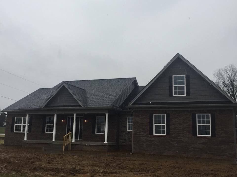 A newly built dark-brick house with a covered front porch, white columns, and a gray shingled roof under a cloudy sky.