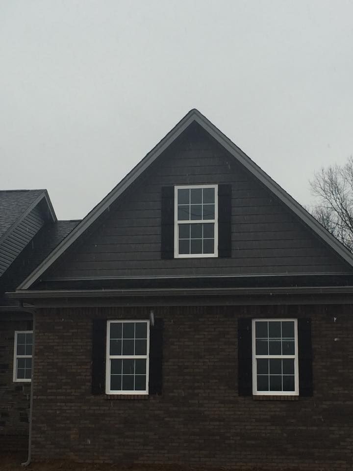 A dark gray gable-roof house exterior with brown brick, black shutters, and three windows under a cloudy sky.