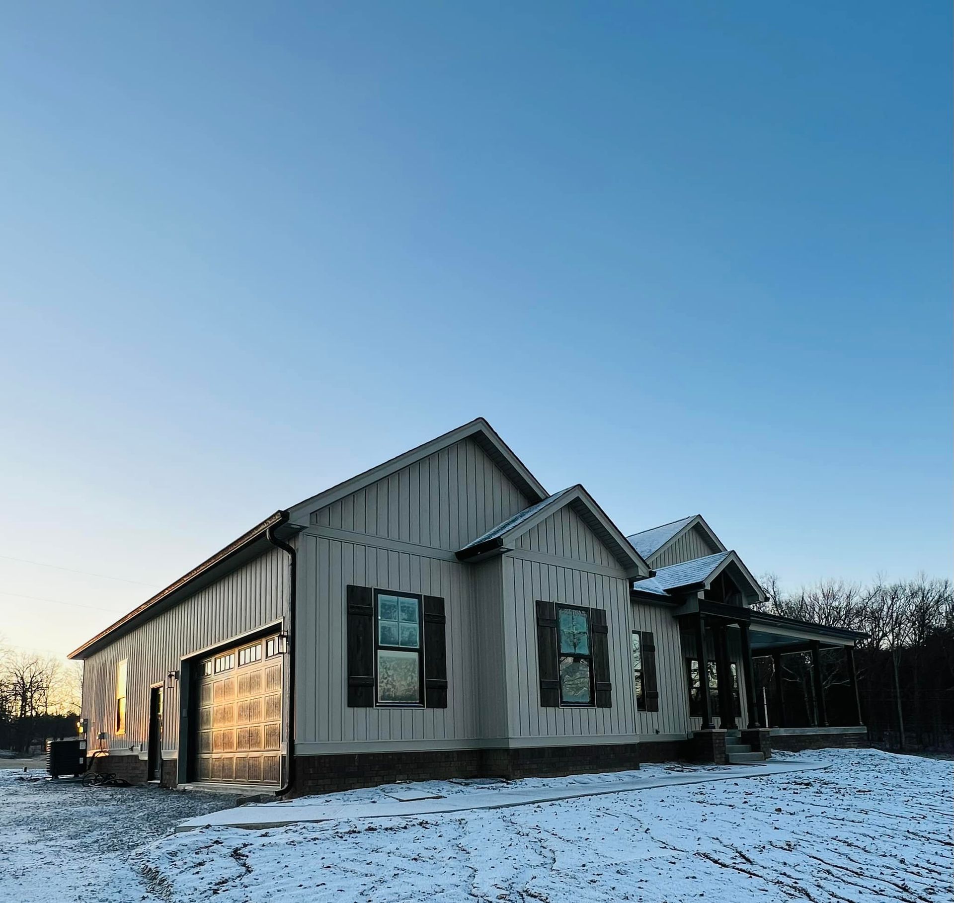A modern light-gray house with dark shutters and a garage stands against a clear blue sky on snow-covered ground.