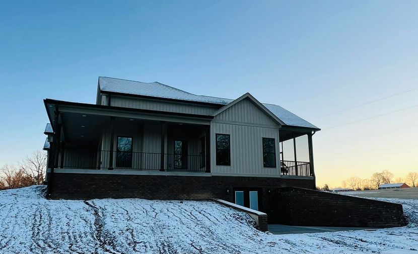 A light gray farmhouse with a large front porch and dark brick foundation stands on a snowy, open landscape at sunrise.