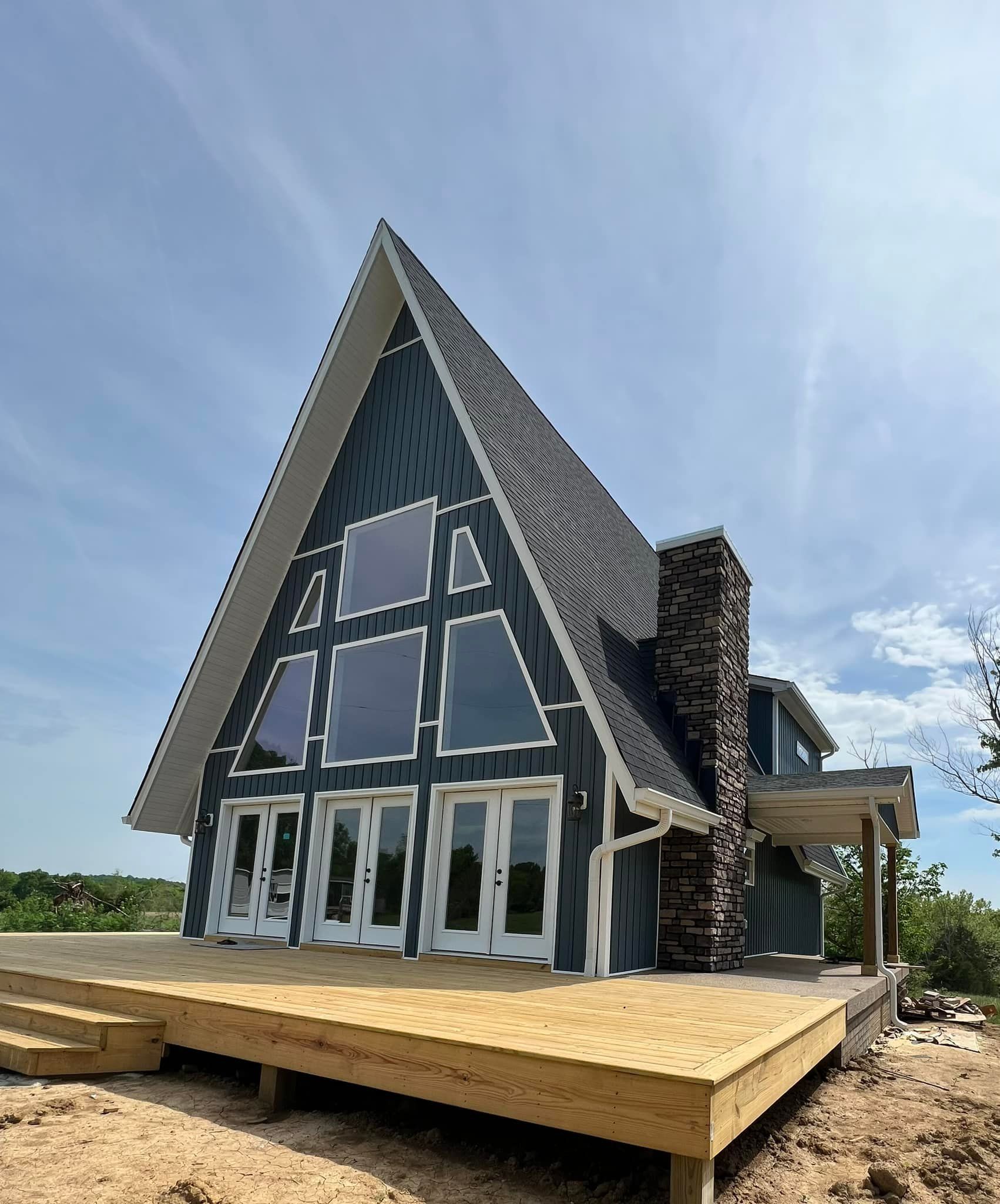 A blue A-frame cabin with a large front deck and stone chimney under a blue, cloudy sky.
