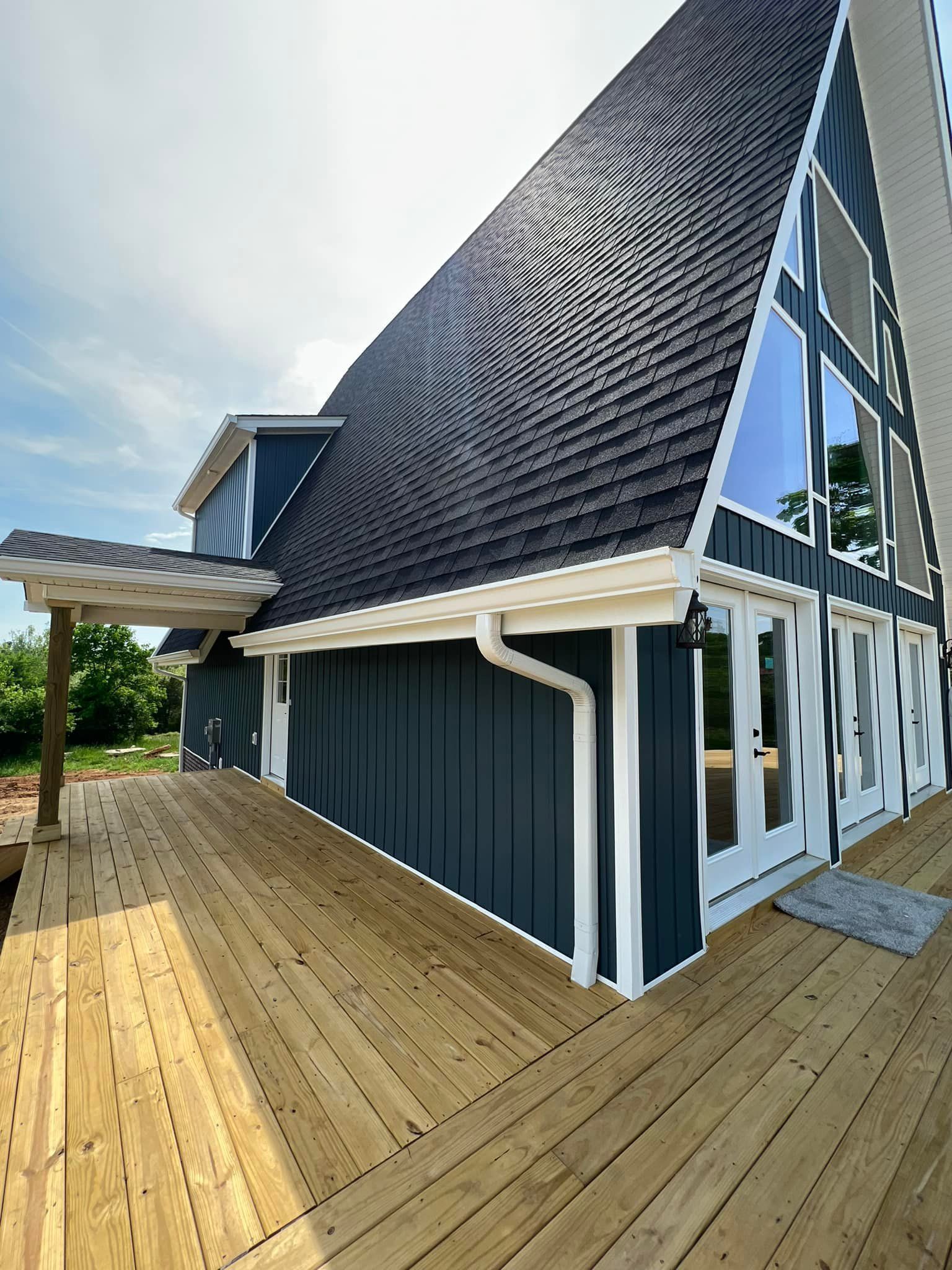 An A-frame house with dark blue vertical siding, a steep shingled roof, and a large wooden deck on a sunny day.
