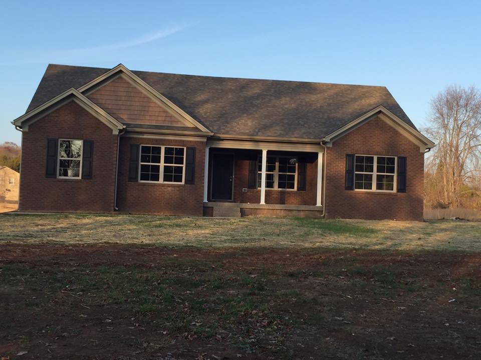 A single-story, brown brick house with a front porch, gabled roof, and dark shutters under a clear blue sky.