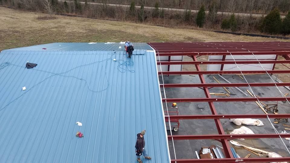 Two people install blue metal roofing panels onto the red steel framework of a building under construction.