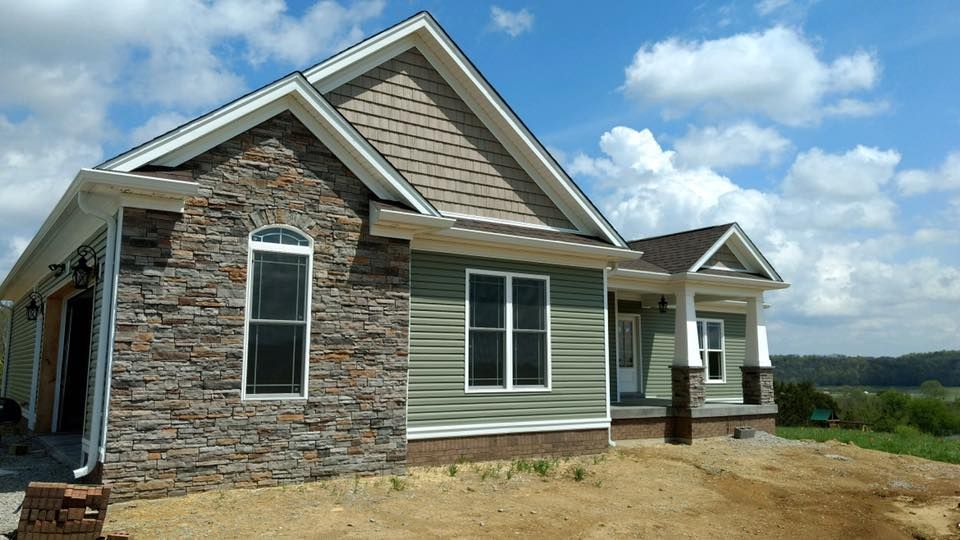 A new single-story house with green horizontal siding, stone facade, and a covered porch under a partly cloudy sky.
