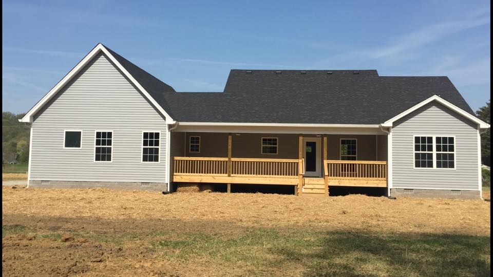A gray, one-story house with a black roof and a wooden deck facing a patch of dirt and grass under a blue sky.