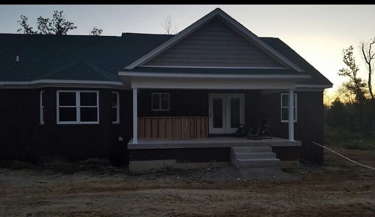 A house under construction with dark siding, a covered porch, white trim, and a dirt yard at dusk.