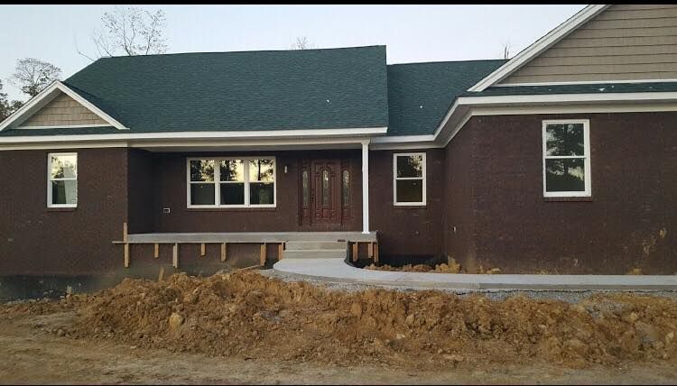 A dark brick single-story house with a green roof, front porch, and unfinished landscaping.