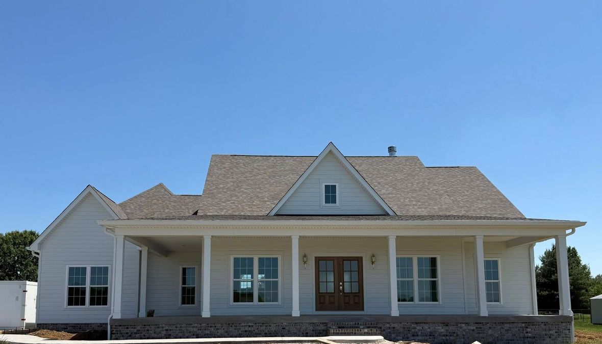 A newly constructed, single-story light gray farmhouse with a covered front porch, white columns, and a brick base.
