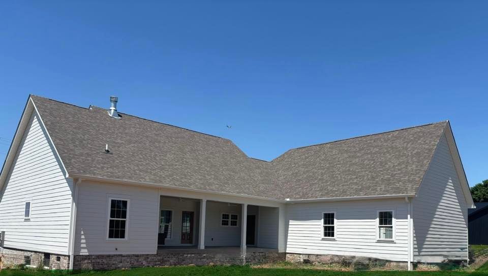 A white, L-shaped single-story house with a grey shingled roof, a covered patio, and a stone foundation against a blue sky.