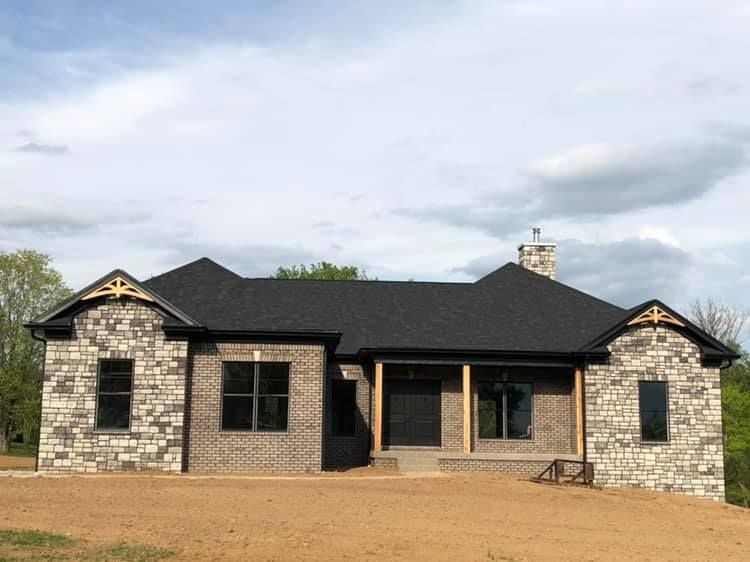 New single-story house with stone and brick exterior, a black shingled roof, and a covered front porch under a cloudy sky.