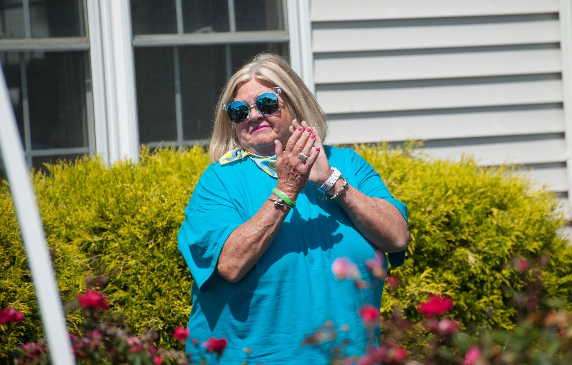 A woman wearing sunglasses and a blue shirt is clapping in front of a house.