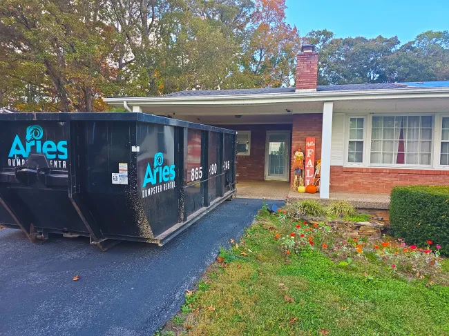 Black dumpster in driveway of a red brick house, likely for a renovation project.